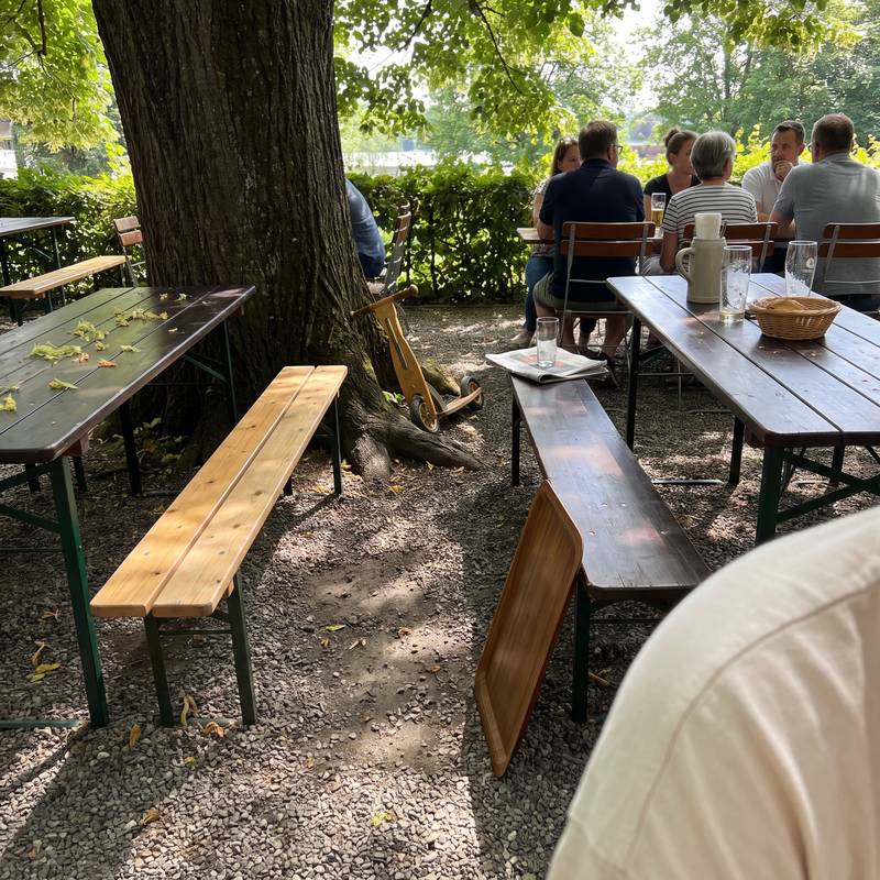 Biergarten am Nachmittag: Biertischgarnituren im Schatten der alten Linde, dahinter G&auml;ste an einem Tisch