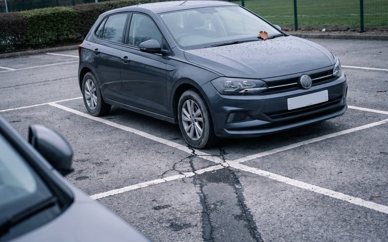 Dunkelgrauer Kompaktwagen mit Schrägheck, von schräg vorn auf einem markierten Stellplatz, Regentropfen auf der Motorhaube, ein Blatt am Scheibenwischer, im Vordergrund unscharf der Kotflügel des Nachbarwagens