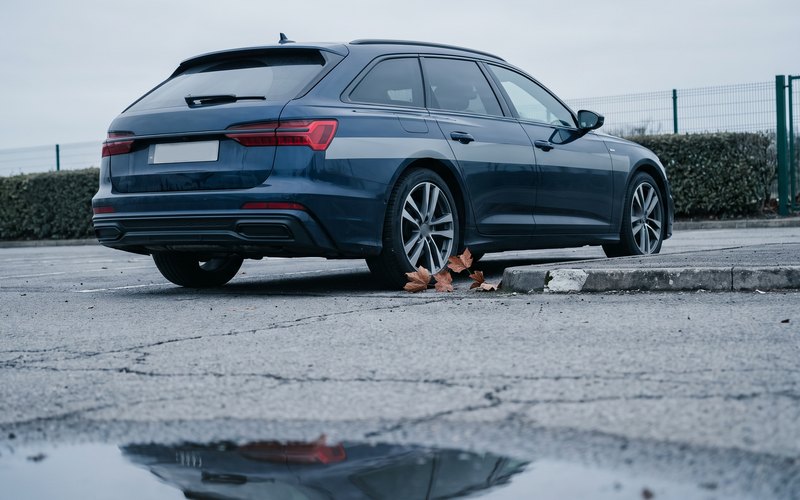 Dunkelblauer Mittelklasse-Kombi von schräg hinten am Bordstein, trockene Blätter am Hinterrad, im Vordergrund unscharf eine flache Pfütze auf dem rissigen Asphalt