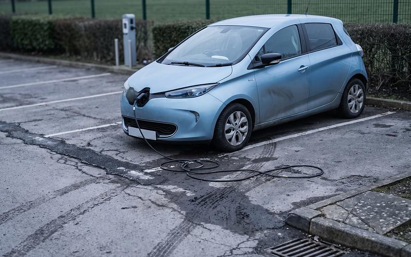 Hellblauer Elektro-Kleinwagen mit offener Ladeklappe an der Front, das Ladekabel steckt und liegt in losen Schlaufen auf dem Asphalt
