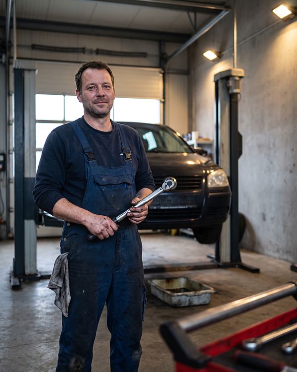 Andreas Kleine in Arbeitskleidung vor der Hebebühne, mit Drehmomentschlüssel in der Hand und Putzlappen in der Beintasche