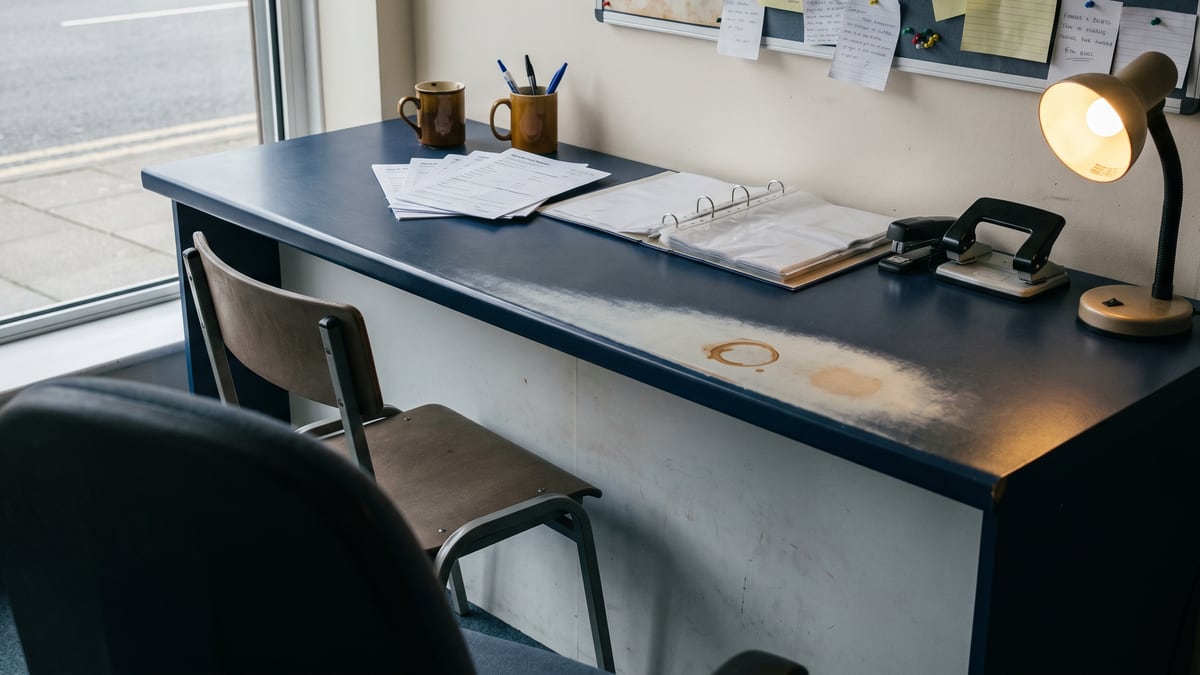 Der leere B&uuml;rotresen mit Anmeldeformularen, einem aufgeschlagenen Ordner, Stiften im Becher und zwei Besucherst&uuml;hlen davor.