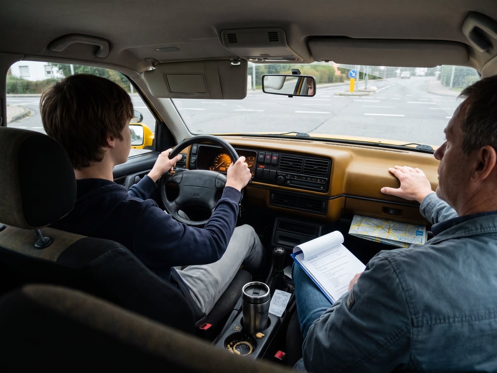 Fahrstunde von der R&uuml;ckbank aus gesehen: eine junge Person am Steuer, daneben der Fahrlehrer mit Klemmbrett auf dem Knie.