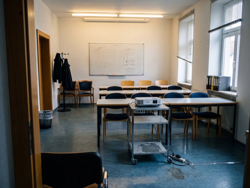 Leerer Schulungsraum &uuml;ber dem B&uuml;ro: Stuhlreihen an schmalen Tischen vor einem Whiteboard, davor ein Beamer auf einem Rollwagen.