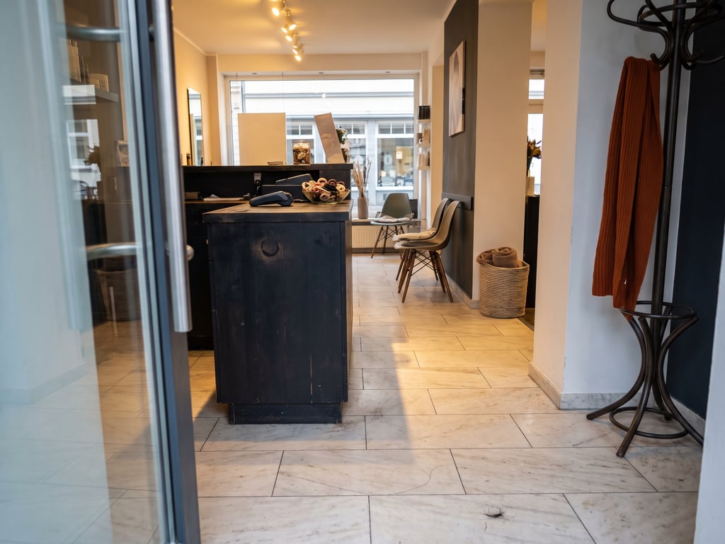 Empfang eines kleinen Friseursalons mit poliertem hellem Marmorboden in großen Platten, dunklem Tresen und Garderobenständer mit Schal