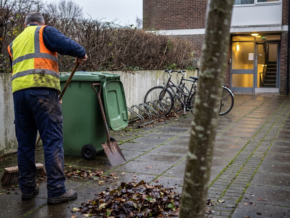 Hausmeister kehrt nasses Laub im gepflasterten Innenhof einer Wohnanlage zusammen, daneben eine offene gr&uuml;ne Tonne