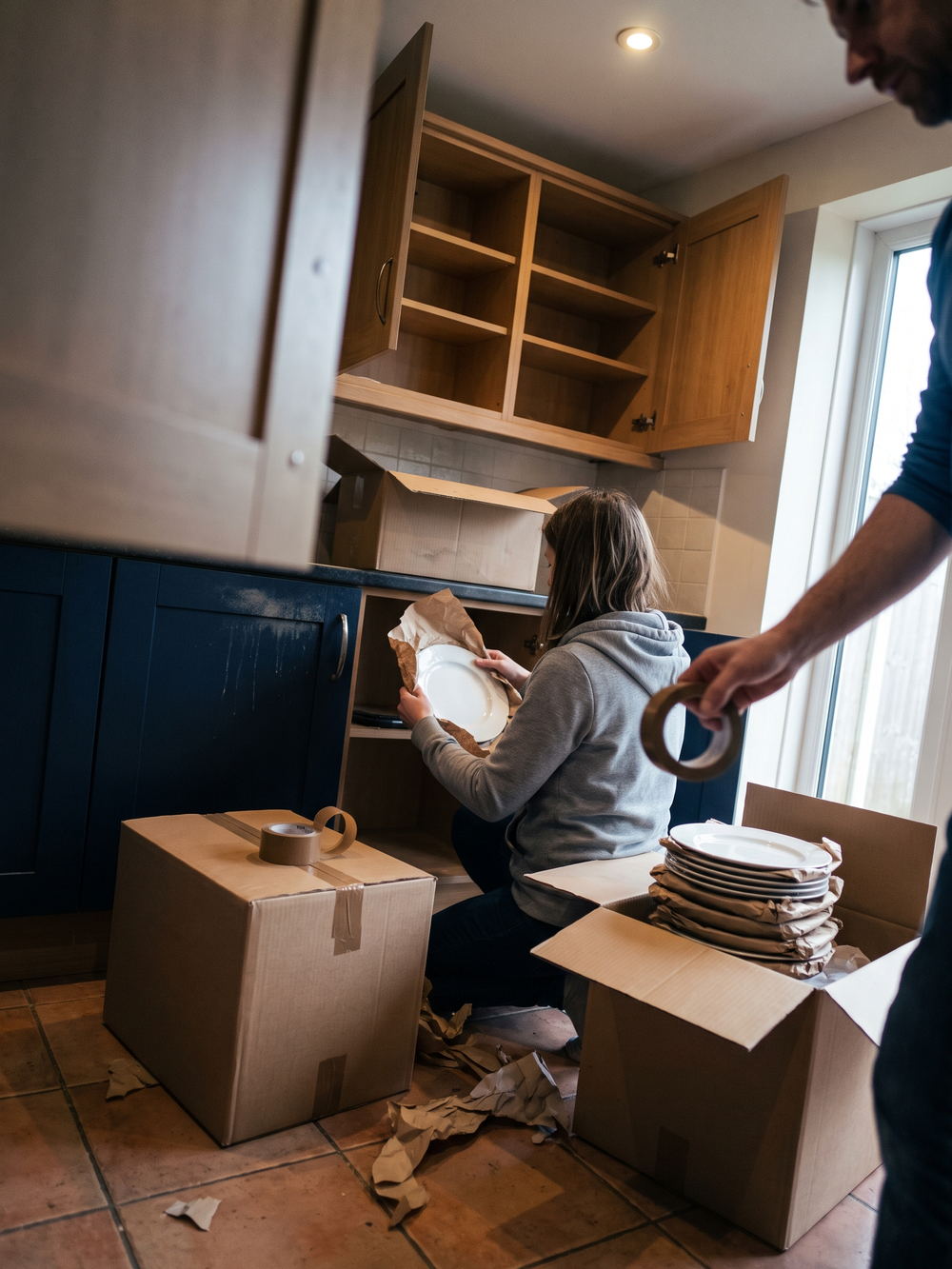 Eine Frau kniet in einer K&uuml;che vor einem ausger&auml;umten Unterschrank und wickelt einen wei&szlig;en Teller in Packpapier; rechts daneben liegen in einem offenen Umzugskarton acht einzeln eingeschlagene Teller flach aufeinander, von rechts reicht ein Unterarm eine Klebebandrolle ins Bild