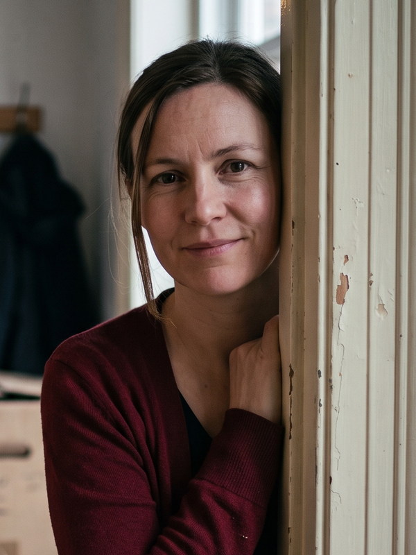 Eine Frau im dunkelroten Cardigan steht am T&uuml;rrahmen einer frisch bezogenen Wohnung und h&auml;lt sich mit einer Hand daran fest, hinter ihr unscharf eine Garderobe mit einem dunklen Mantel