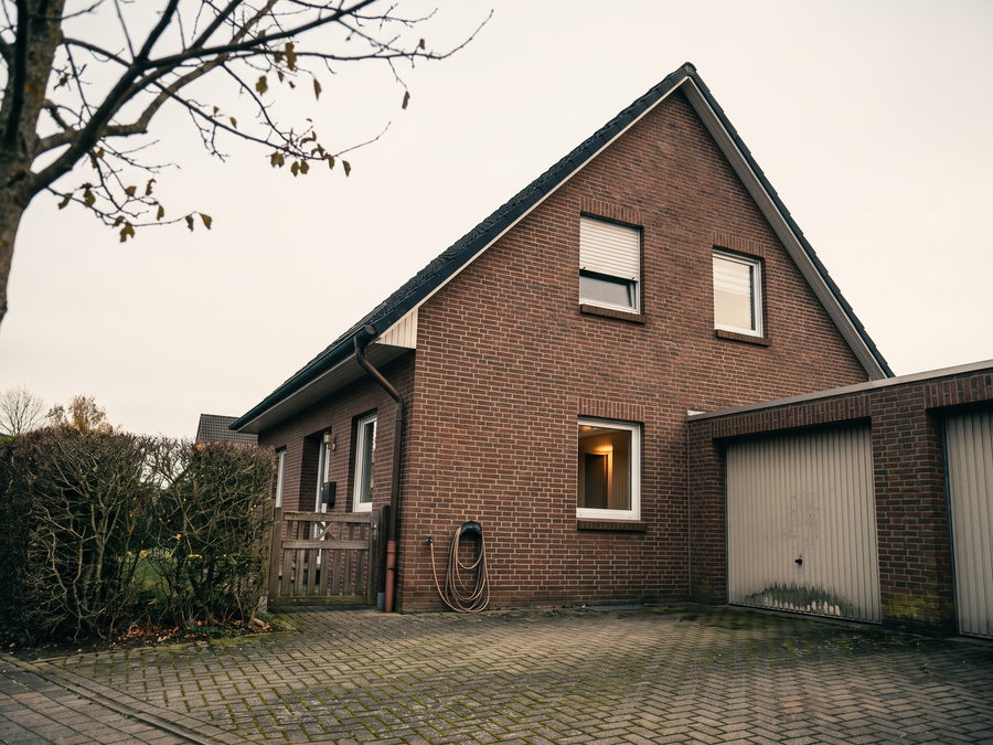 Freistehendes Einfamilienhaus aus Klinker mit Satteldach, rechts das geschlossene Garagentor, davor eine gepflasterte Einfahrt mit Moos in den Fugen