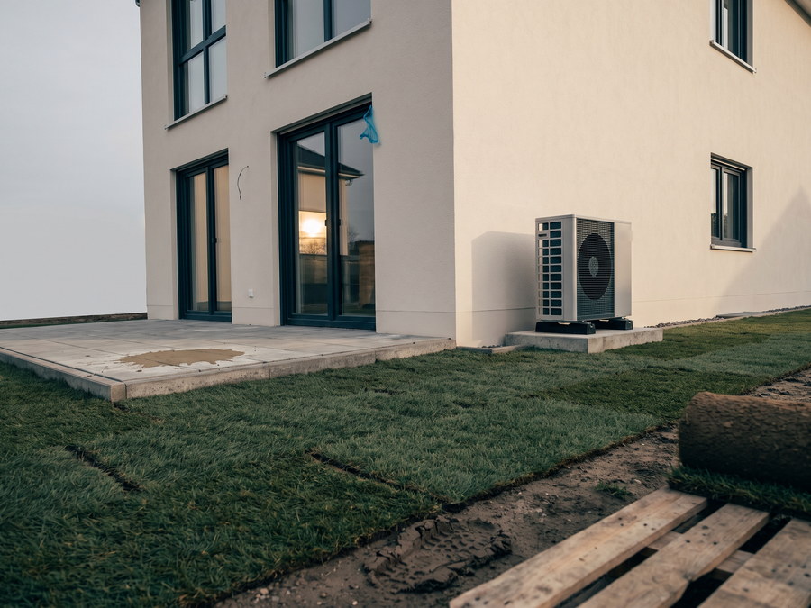 Gartenseite einer Neubau-Doppelhaush&auml;lfte mit hellem Putz und gro&szlig;en Fenstern, neben der Hauswand eine W&auml;rmepumpe auf einem Betonsockel, im Vordergrund frisch verlegter Rollrasen mit sichtbaren N&auml;hten