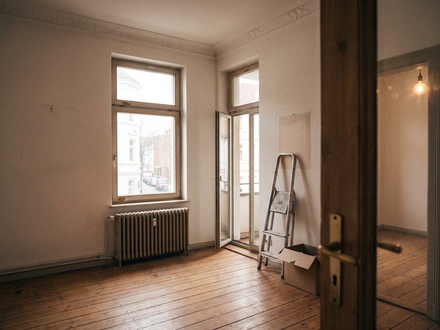 Leeres Altbauzimmer mit Dielenboden, Stuckleiste an der Decke, hohem Fenster und gusseisernem Heizk&ouml;rper, daneben eine halb offene Balkont&uuml;r und eine zusammengeklappte Trittleiter