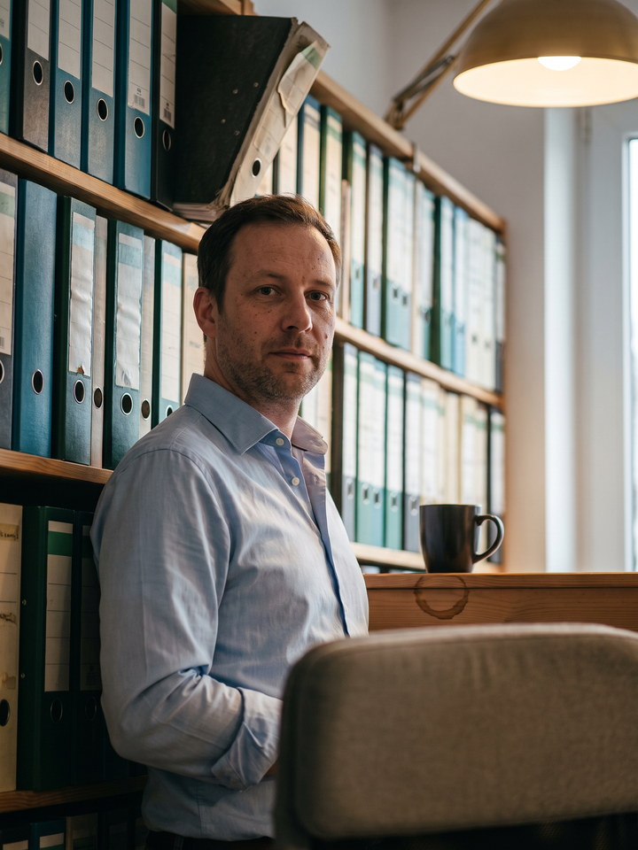 Andreas Vogt im hellen Hemd vor einer Regalwand mit Ordnern, auf dem Regalbrett eine Kaffeetasse