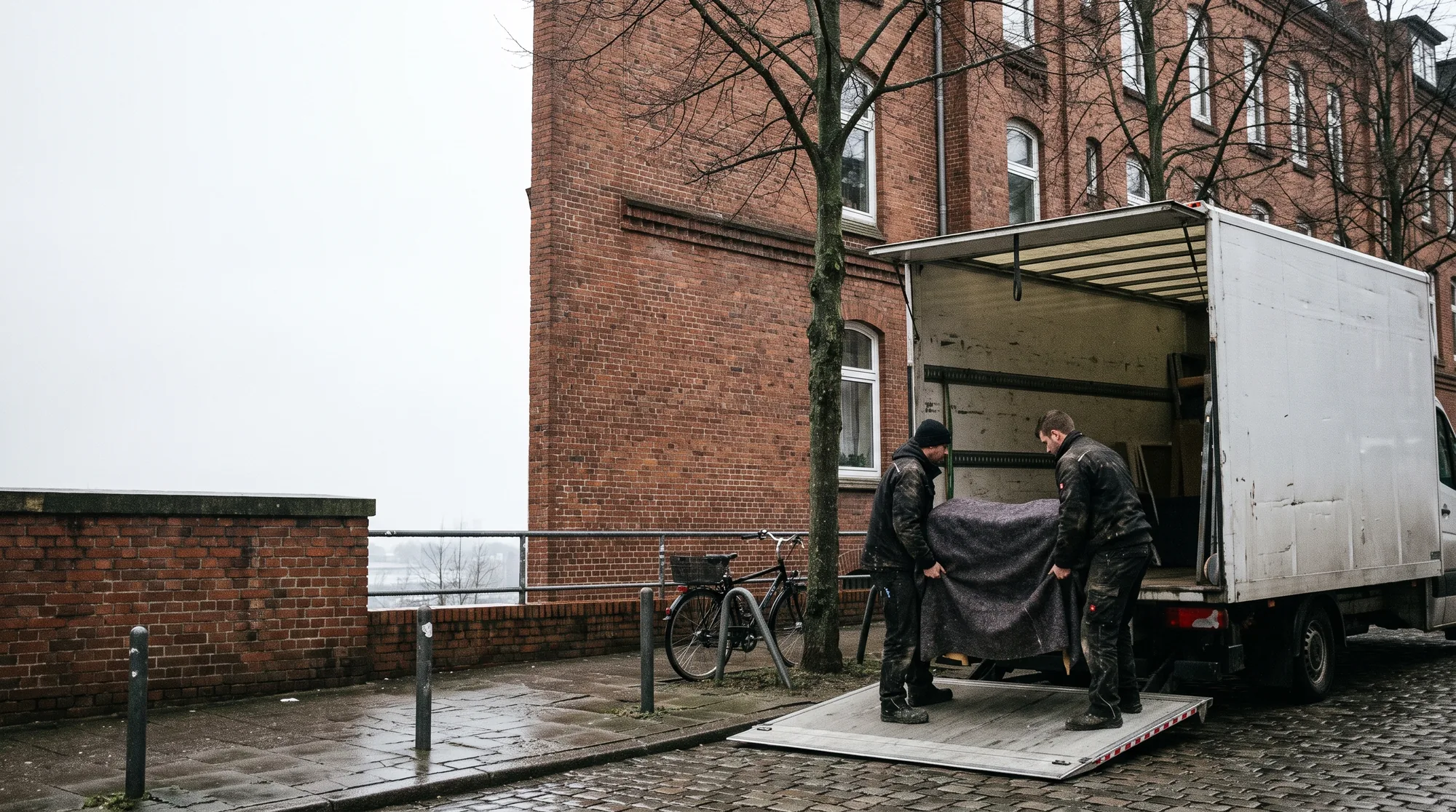 Ein weißer Umzugswagen am Bordstein einer Hamburger Straße, die Ladebordwand abgesenkt, zwei Möbelpacker tragen ein verpacktes Möbelstück über die Rampe, dahinter ein Backsteinaltbau.
