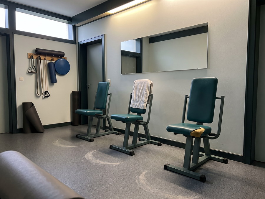 Trainingsraum mit drei Sequenztrainingsger&auml;ten in dunklem Gr&uuml;n mit petrolfarbenen Polstern, im Gummiboden davor zwei hell abgelaufene Bogen, an der Wand ein Regal mit B&auml;ndern, Faszienrolle und Balancekissen