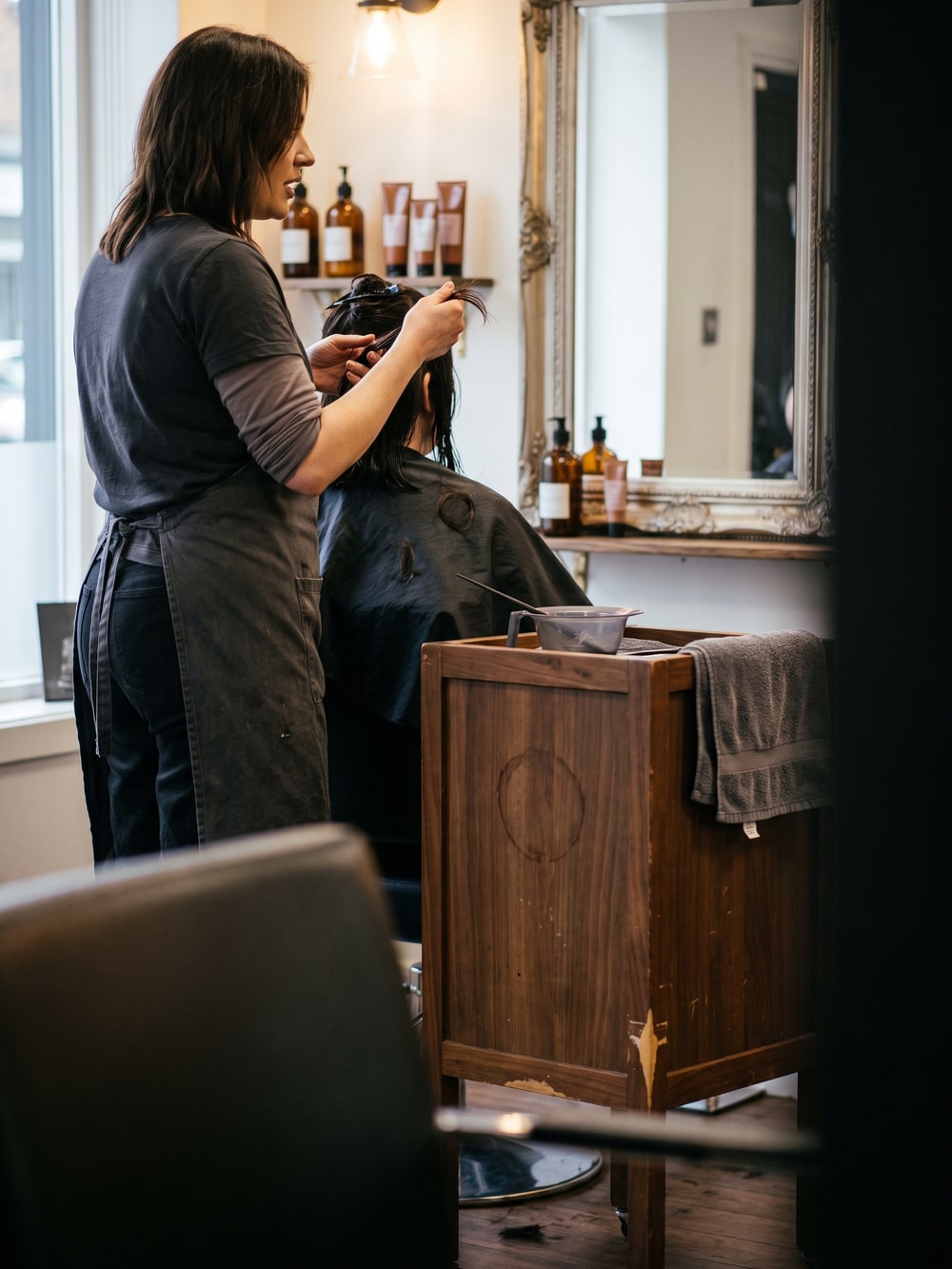 Friseurin hält am Spiegelplatz eine Haarsträhne der sitzenden Kundin zwischen zwei Fingern hoch, daneben ein Rollwagen mit Färbeschale und Pinsel