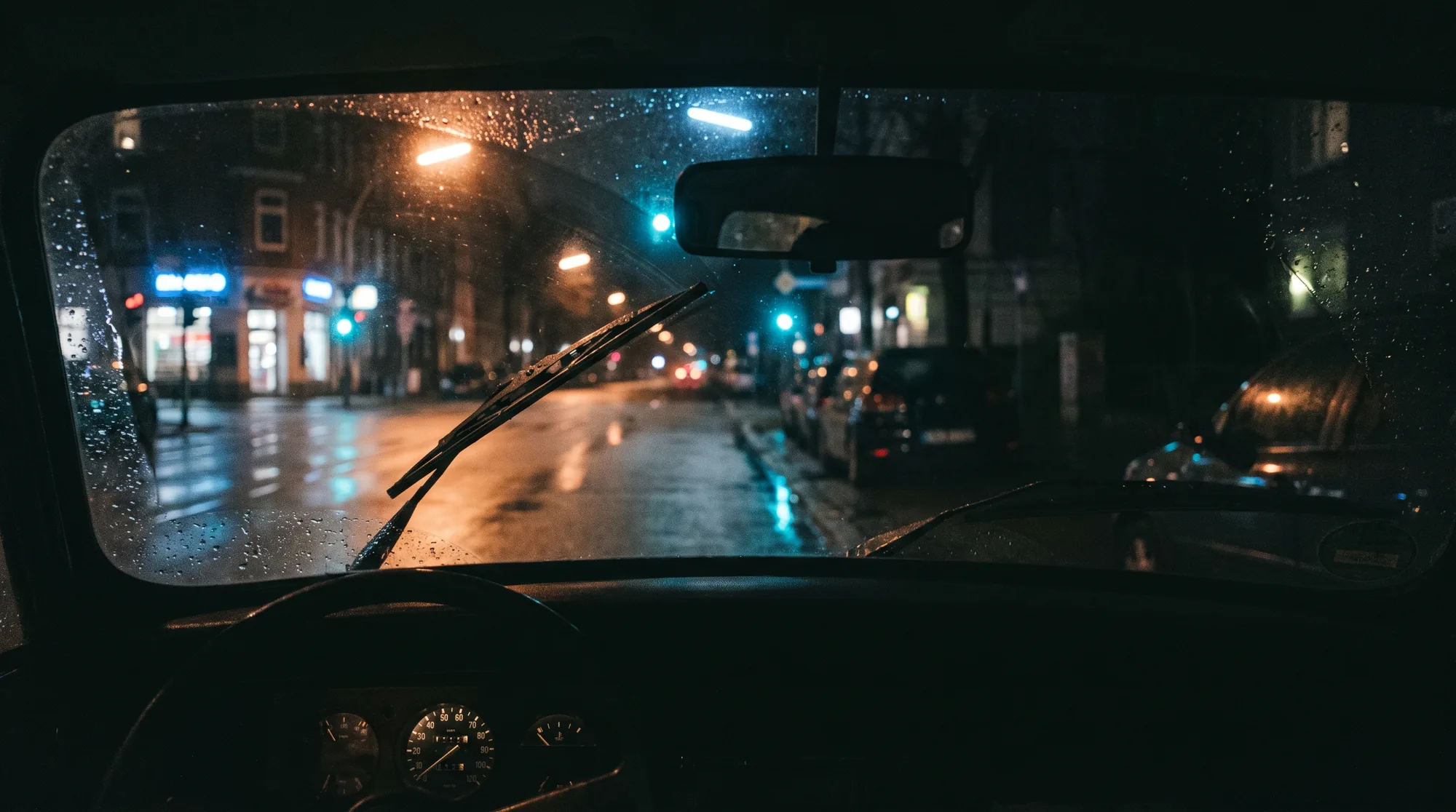 Blick durch die Windschutzscheibe eines Fahrschulwagens bei Nacht auf eine nasse Hamburger Straße, Ampeln und Ladenlichter spiegeln sich im Asphalt.