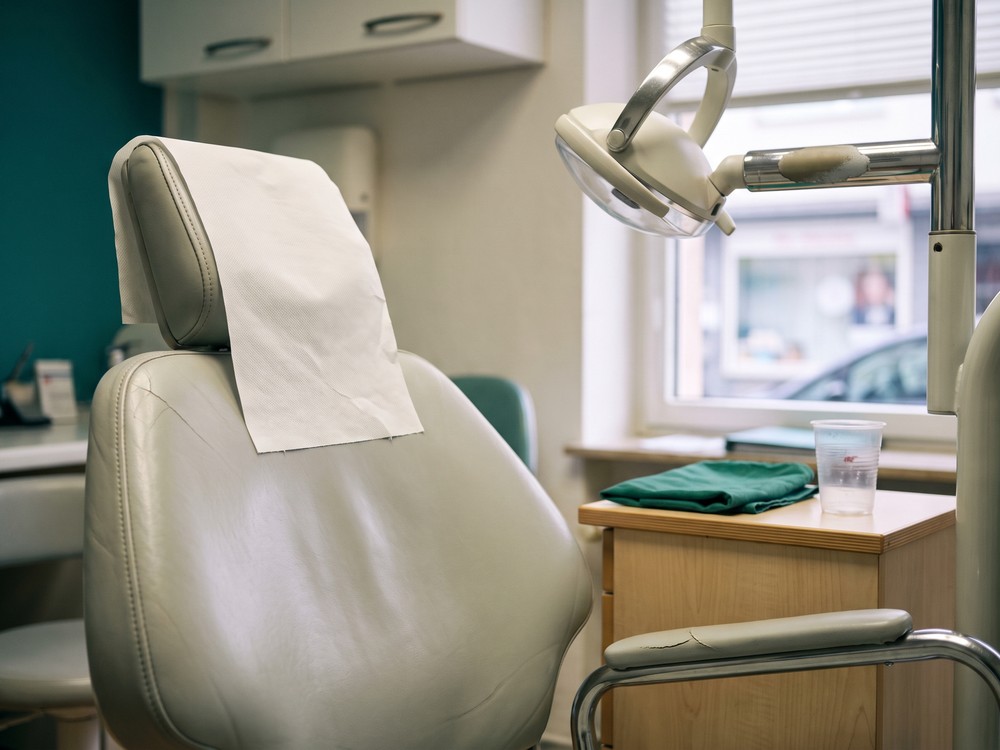 Behandlungsstuhl aus der Sicht des Patienten, daneben ein Beistelltisch mit gefaltetem gr&uuml;nem Tuch und einem Sp&uuml;lbecher; die Behandlungsleuchte ist ausgeschaltet zur Seite geschwenkt