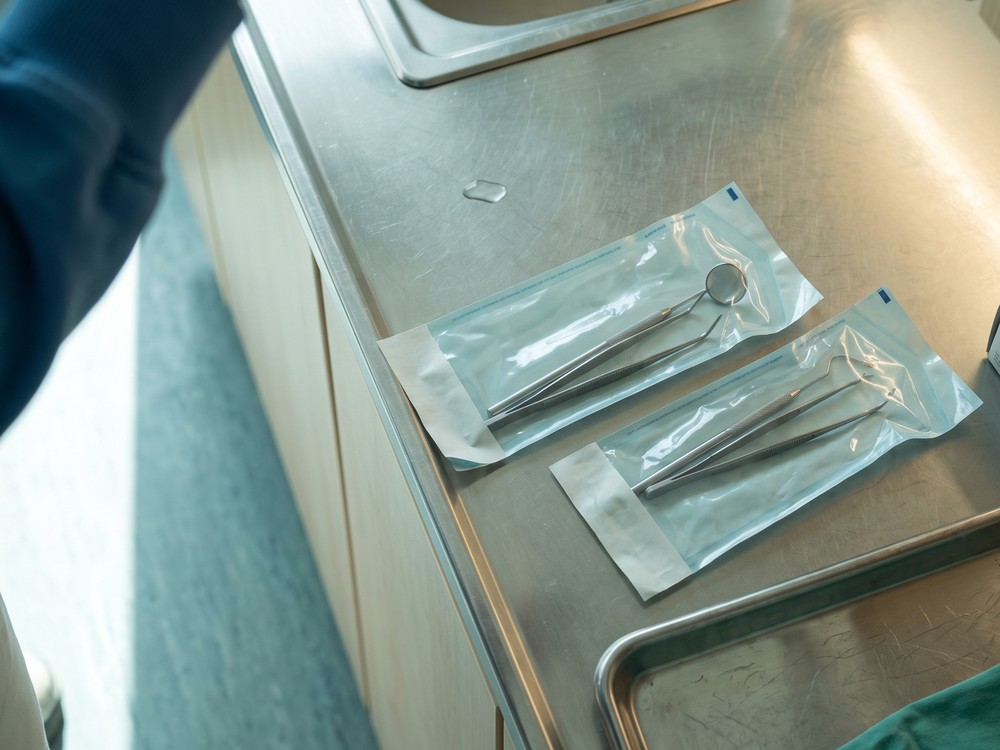 Zwei verschwei&szlig;te Sterilbeutel mit Mundspiegel, Sonde und Pinzette auf der Edelstahl-Arbeitsplatte neben dem Becken