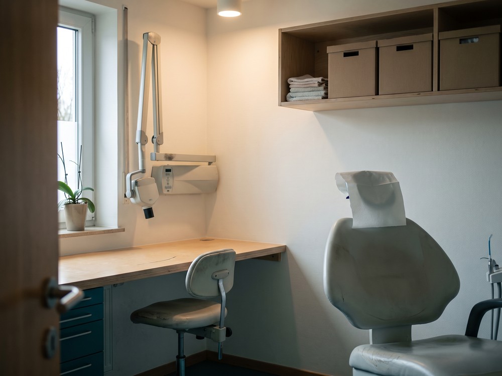 Behandlungszimmer mit Tageslicht: der Behandlungsstuhl mit frischer Papierauflage an der Kopfst&uuml;tze, davor ein Rollhocker am Arbeitstresen, links das Fenster mit einer Pflanze auf der Bank