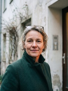 Marion Kirchner mit kurzem grauem Haar und hochgeschobener Lesebrille vor einer Haust&uuml;r